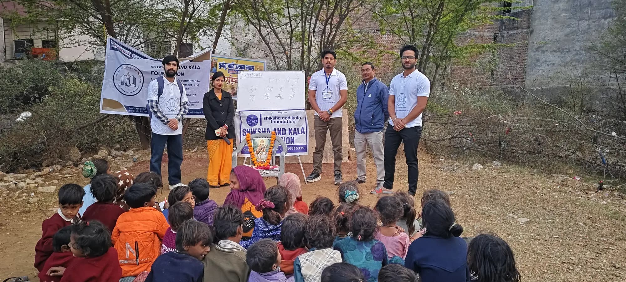 Children attending an outdoor Hindi-vowel lesson beside a foundation banner and learning board.