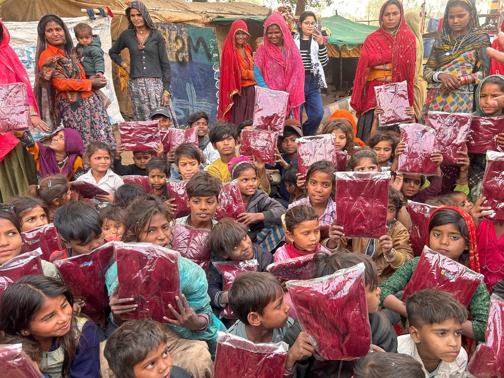 Children and community members holding packaged sweaters during winter support at Rajawas Kalbeliya Basti.
